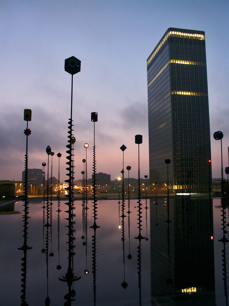 Mehrere Pfähle und Gebäude mit Lichtern unter einem bewölkten Himmel, mit Spiegelungen der Pfähle.