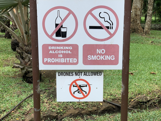 Schild am Straßrand, das das Trinken von Alkohol und Rauchen verbietet, mit Bäumen und Gras im Hintergrund.