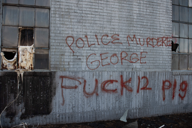 Ein Gebäude mit der Graffiti-Beschriftung "Polizei ermordet George Fuck 12 P19" an der Seite, mit Fenstern und diversen Gegenständen vorne.