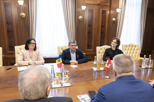 Eine Gruppe von Menschen sitzt um einen Holztisch in einer formellen Umgebung und ist in eine Diskussion mit EU-bezogenen Materialien auf dem Tisch vertieft, einschließlich Flaggen, Papieren und mobilen Geräten.