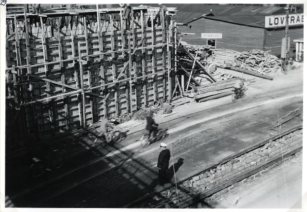 Schwarz-weiß-Foto einer Baustelle mit einem teilweise errichteten Gebäude, Menschen, die Fahrräder fahren und gehen, Bahnschienen, Strommasten, Holzplanken und Text am unteren Rand.