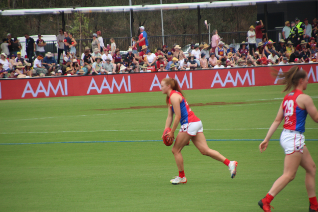 Zwei Frauen beim Australian Football League-Spiel auf einem Feld, eine hält den Ball, mit Zuschauern, Tafeln, Zäunen, Pfosten und Bäumen im Hintergrund.