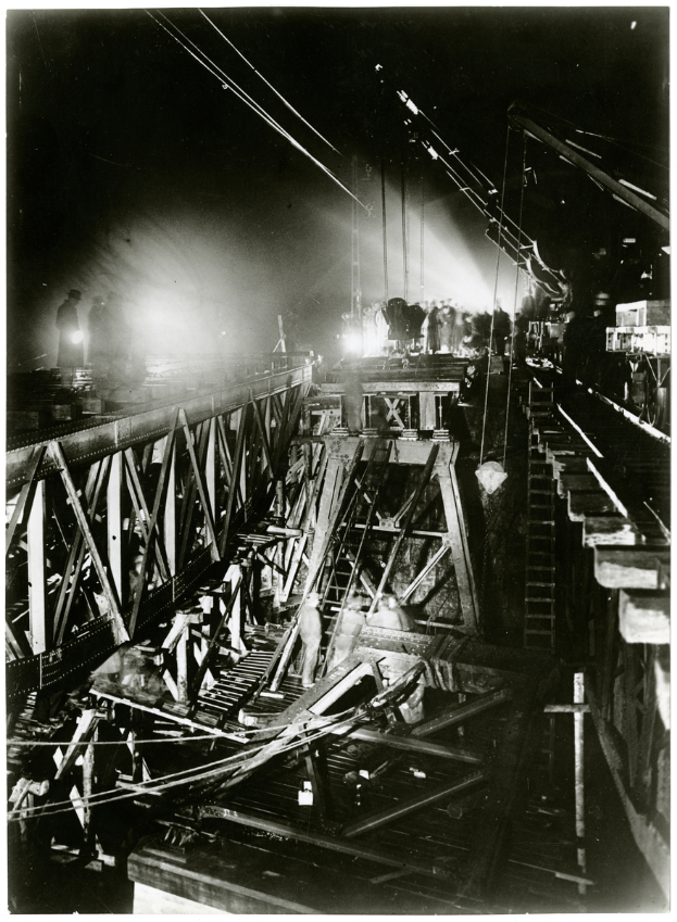 Ein Schwarz-Weiß-Foto einer Brücke im Bau, mit ein paar Menschen auf der Brücke und ein paar Lichtern, die den Bereich beleuchten.