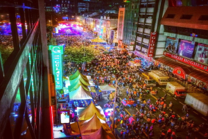 Ein lebhaftes Karneval-Szenario mit einer großen Menschenmenge auf der Straße, umgeben von Zelten, Geschäften, Gebäuden und Bannern, mit zahlreichen Lichtern, die den Hintergrundbereich erhellen.