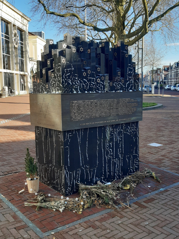 Denkmal in einem Stadtplatz zu Ehren von Holocaust-Opfern mit einem Baum davor, einer Topfpflanze auf dem Boden, verstreuten trockenen Blättern und Gebäuden, Fahrzeugen und Bäumen im Hintergrund.
