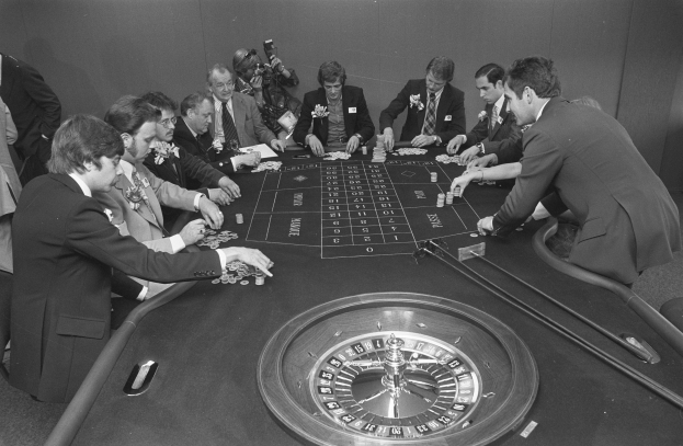 Eine Gruppe von Männern in Blazern und Krawatten sitzt um einen Roulettetisch in einem Casino, hält Karten und ist von Münzen und anderen Gegenständen umgeben, mit einer Wand im Hintergrund, dargestellt in Schwarz-Weiß.