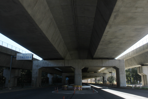 Autobahn unter einer Brücke mit Absperrgittern, Pollern und Texttafeln am Straßenrand, Geländer an den Seiten, Bäume und einen klaren blauen Himmel im Hintergrund.