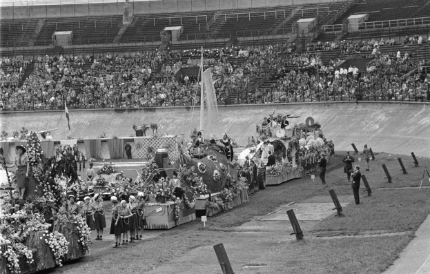 Schwarzes und weißes Foto eines Umzugs in einem Stadion mit Menschen, die stehen und sitzen, einer zentralen Fontäne und Blumensträußen auf Fahrzeugen.