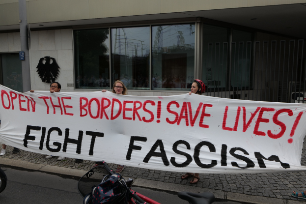 Gruppe von Menschen mit einem Transparent 'Öffnet die Grenzen, rettet Leben, bekämpft Faschismus' vor einem Gebäude mit Glasfenstern und einer Stange, mit einem Fahrrad und einer Tasche auf der Straße.