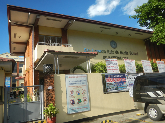 Ein Schulgebäude mit einem vor dem Gebäude geparkten Van, das Fenster, Geländer, Säulen, ein Dach, Banner mit Text, Topfpflanzen, ein Metalltor, eine Gruppe von Bäumen und einen bewölkten Himmel im Hintergrund zeigt.