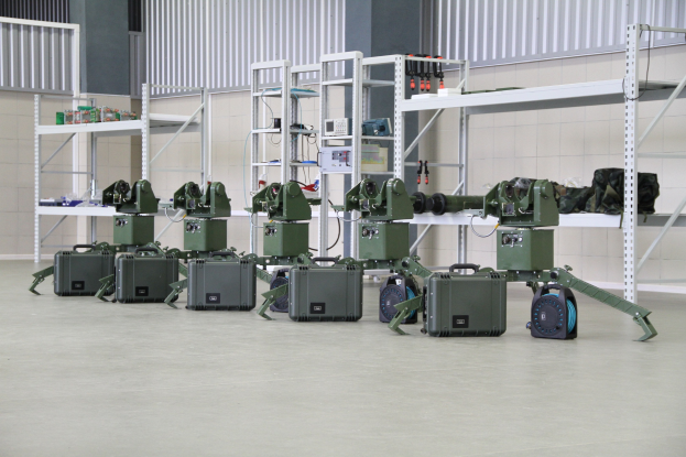 Eine Gruppe von Milit├Ąrrobotern auf einem Boden mit Koffern und Objekten auf Gestellen, die in der N├Ąhe einer Wand und eines Gel├Ąnders als Teil eines Verteidigungssystems positioniert sind.