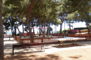 Eine Parkszene mit einem zentralen Tischtennis-Tisch, umgeben von Bänken, einem Zaun, Schaukeln, Pfählen, Pflanzen und Bäumen, mit einem Wasserlauf und einem klaren blauen Himmel im Hintergrund.