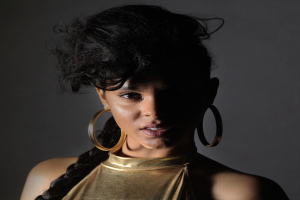 Eine Frau in einem goldenen Kleid und großen Ohrringen steht selbstbewusst vor einer Wand, ihr Haar ist zu einem sleeken Bob gestylt und sie schaut direkt in die Kamera mit einem entschlossenen Gesichtsausdruck.