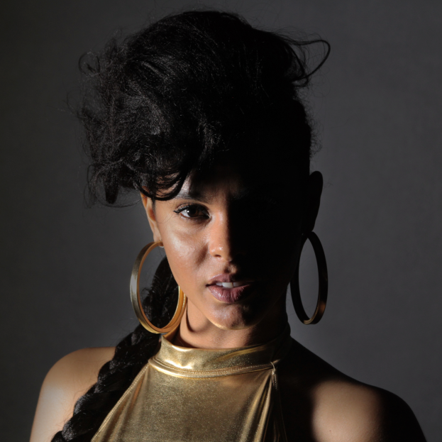 Eine Frau in einem goldenen Kleid und großen Ohrringen steht selbstbewusst vor einer Wand, ihr Haar ist zu einem sleeken Bob gestylt und sie schaut direkt in die Kamera mit einem entschlossenen Gesichtsausdruck.