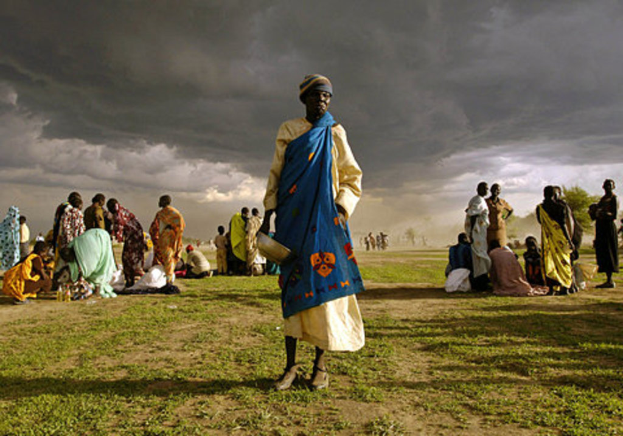 Eine Frau in einem blauen Kleid steht mit einem silbernen Gefäß in der Hand und wird von einer Gruppe Frauen umgeben, die hinter ihr sitzen oder stehen, unter einem bewölkten Himmel mit Bäumen im Hintergrund.
