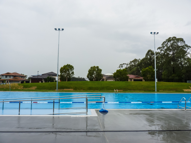 Großer rechteckiger Schwimmbad im Park, umgeben von Zäunen und Pfählen mit Überdachungsleuchten, umgeben von Bäumen, mit Häusern und einem klaren blauen Himmel im Hintergrund.