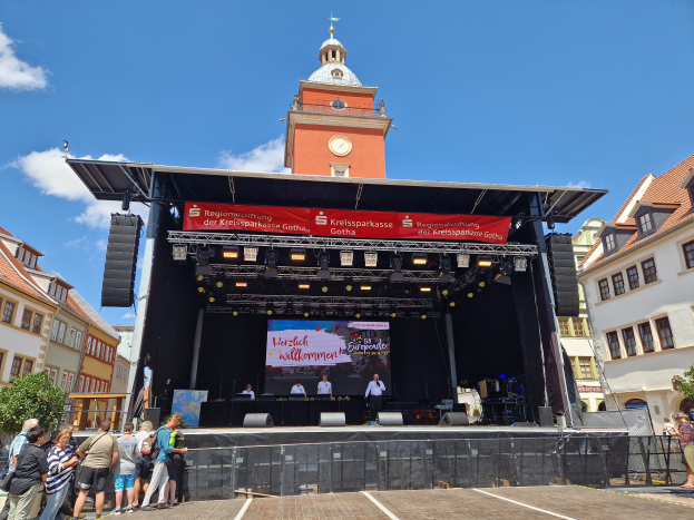 Eine Gruppe von Menschen auf einer Bühne vor einem Uhrturm, umgeben von Gebäuden, Bäumen und einem bewölkten Himmel, mit einem Bildschirm auf der Bühne, beim Kreissparkasse Gotha Musikfestival.