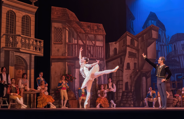 Zwei Tänzer performen auf einer Bühne im Royal Opera House in London, mit einer projizierten Stadtlandschaft als Hintergrund und einem Publikum, das sowohl sitzt als auch steht.