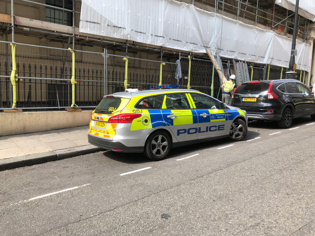 Ein Polizeiwagen steht auf der Seite einer Straße neben einem Gebäude, daneben stehen zwei Helme tragende Personen; im Vordergrund sind ein Geländer und ein Pfahl zu sehen, im Hintergrund hängt eine weiße Decke über das Gebäude.
