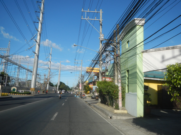 Stadtstraße mit Stromleitungen, fahrenden Fahrzeugen, Gebäuden, Bäumen, Schildern, Laternen und fernen Türmen.