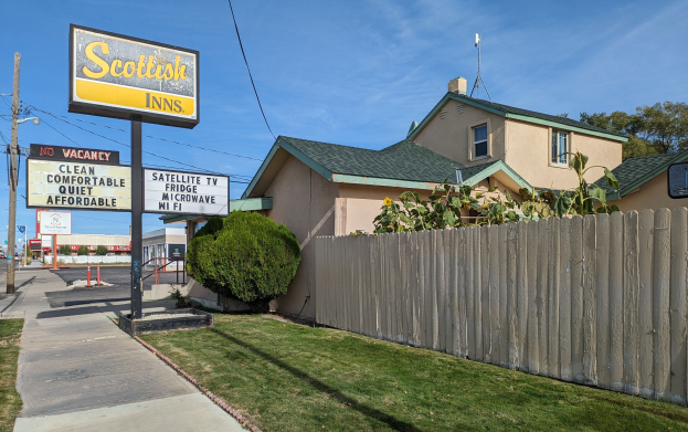 Ein schottisches Motel in San Francisco, Kalifornien, mit einem Haus mit Dach und Fenstern, einem hölzernen Zaun, Pflanzen, Gras, einer Gruppe von Bäumen, Schildern, Strommasten mit Drähten, einer Straßenlaterne, Fahrzeugen auf der Straße und einer bewölkten Himmel.