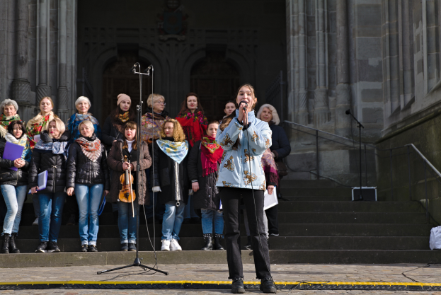 Eine Gruppe von Menschen steht vor einem Gebäude, eine Frau in der Mitte hält ein Mikrofon und singt, umgeben von Mikrofonen mit Ständern, Musikinstrumenten, Treppen mit Geländern, einer Wand mit Säulen und einer Tür auf der rechten Seite.