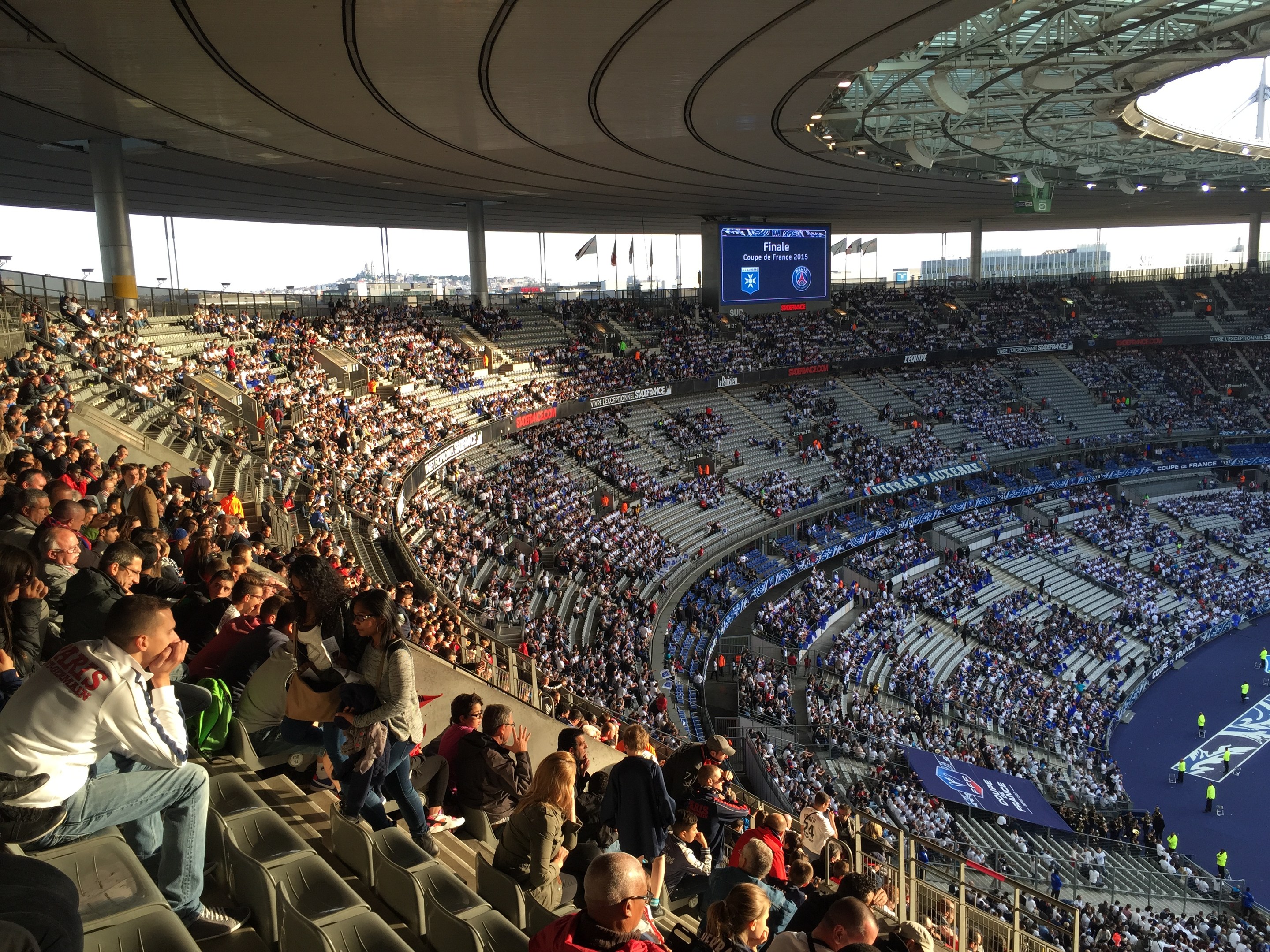 Große Menschenmenge in einem Stadion bei einem Fürballspiel mit einer Bühne, Fahnen, Masten und einem Bildschirm im Hintergrund.