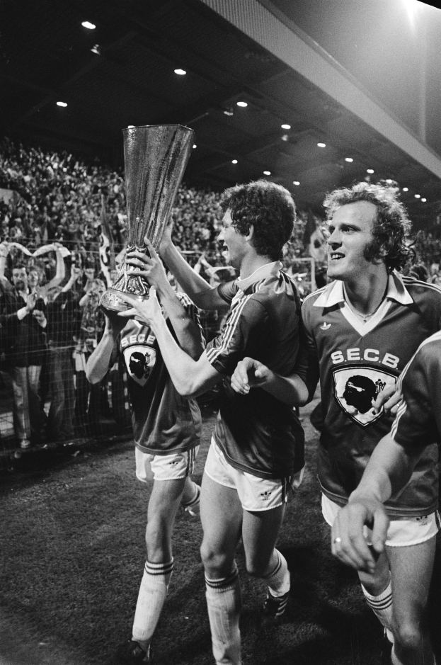 Schwarzes Bild von Fußballspielern, die mit einem Pokal auf einem Feld feiern, umgeben von einer Menge hinter einem Zaun unter Stadionlichtern.