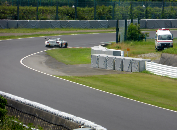 Ein Rennwagen fährt eine Rennstrecke entlang, im Hintergrund ist ein Polizeiwagen zu sehen, umgeben von Gras, Pflanzen, Bäumen und einem Zaun, mit einem Fahrzeug auf der rechten Seite und Text unten.
