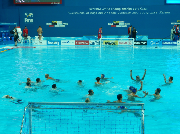 Eine Gruppe von Menschen, die Wasserball in einem Pool mit einem Netz spielen, mit Zuschauern und Tafeln mit dem Text "FINA World Championships 2015 Kazan" im Hintergrund.