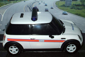 Weißer Polizeiwagen vor bewegendem Verkehr auf einer Straße, die mit Gras und Bäumen gesäumt ist.