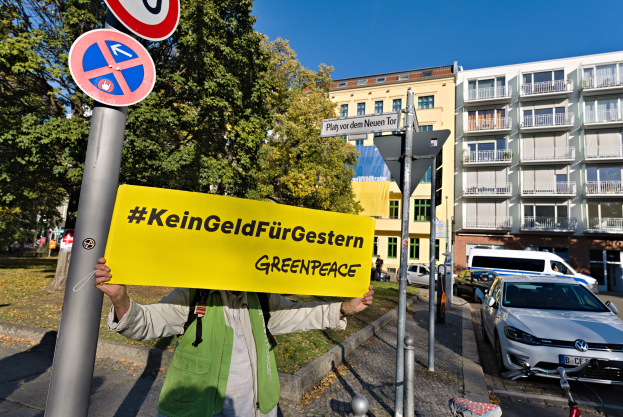 Eine Person mit einer Tasche und einem "Greenpeace"-Schild vor einem Gebäude mit Fahrzeugen, einem Fahrrad und Schildern auf Pfählen, mit Bäumen und einem klaren blauen Himmel im Hintergrund.