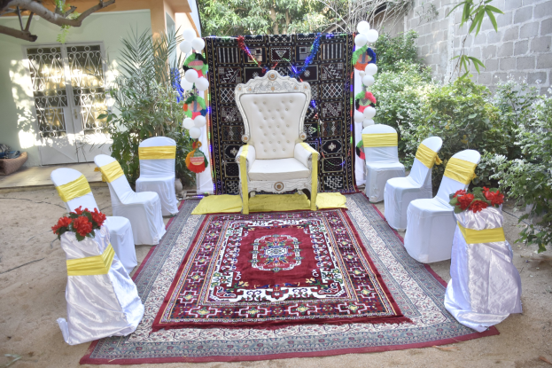 Eine Gartenhochzeitsaufstellung mit weißen Stühlen, einem roten Teppichläufer, Blumenbouquets, Dekorationsballons, Topfpflanzen und einem Gebäude mit Fenstern im Hintergrund.