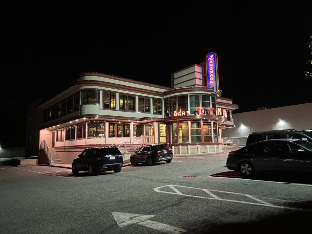 Restaurant bei Nacht mit parkenden Autos, Glasfenstern, Treppen mit Geländern, beleuchteter Bereich, Namensschild und einem Baum auf der rechten Seite in einer dunklen, mysteriösen Umgebung.