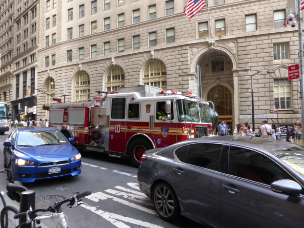 Ein Feuerwehrauto parkt am Straßenrand in New York City, mit Fahrzeugen auf der Straße, Passanten auf dem Gehweg und Fahrrädern in der Nähe, mit Gebäuden, Laternenmasten und Fahnenmasten im Hintergrund.