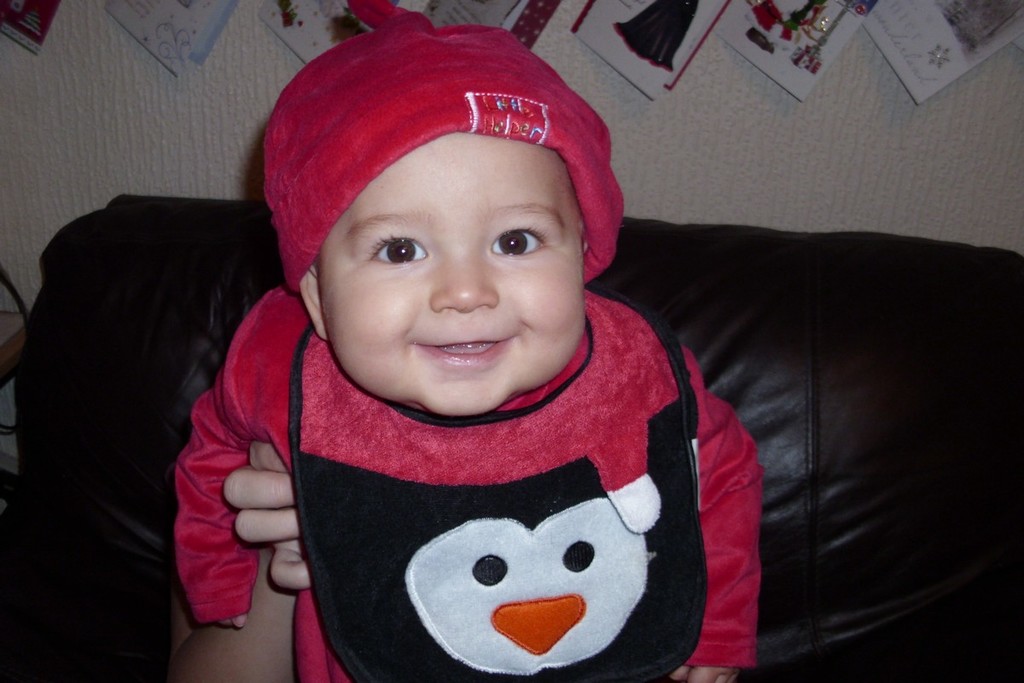 Ein Baby in einem roten Kleid und Hut sitzt auf einem braunen Sofa mit Wandkarten im Hintergrund.