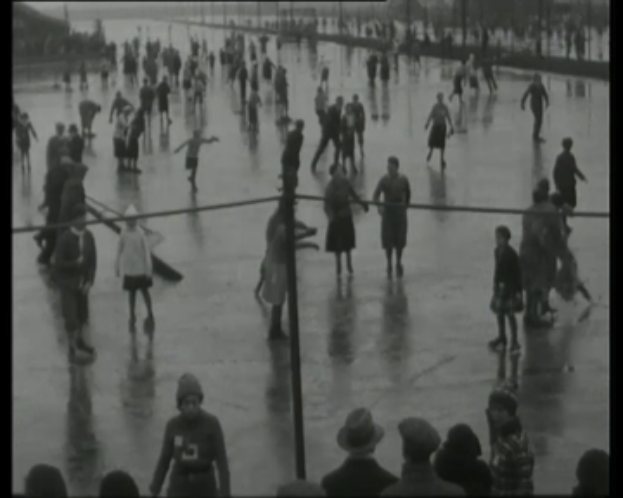 Eine Gruppe von Menschen, die auf einer Eisbahn Schlittschuh laufen, bei Regen, mit Pfählen und Bäumen im Hintergrund, dargestellt in Schwarz-Weiß.