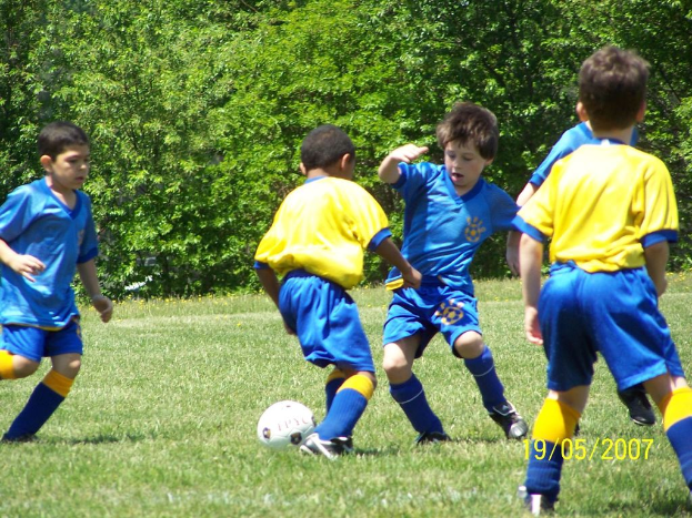 Kinder spielen Fußball auf einem grünen Feld mit Bäumen im Hintergrund und einem Datum in der rechten unteren Ecke.