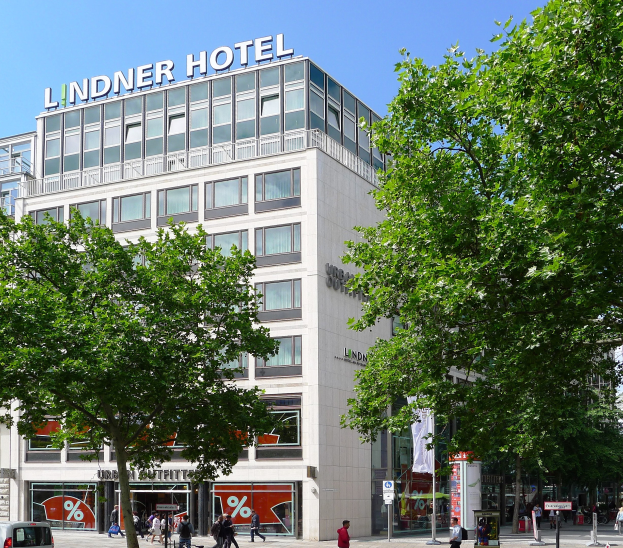 Großes Glasfrontgebäude mit "Lindner Hotel"-Text oben, umgeben von Bäumen und einer belebten Straße mit Fußgängern und Fahrzeugen.