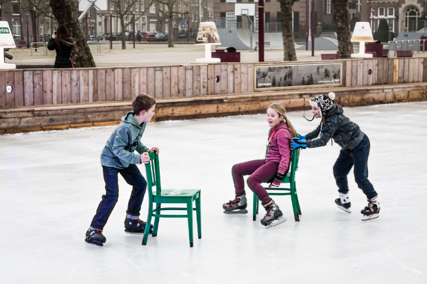 Kinder beim Skifahren im Vordergrund eines Spielplatzes mit drei Kindern und zwei St"hlen in der Mitte und Geb"uden, B"umen, B"anken, Pfosten und einem Basketballfeld im Hintergrund.