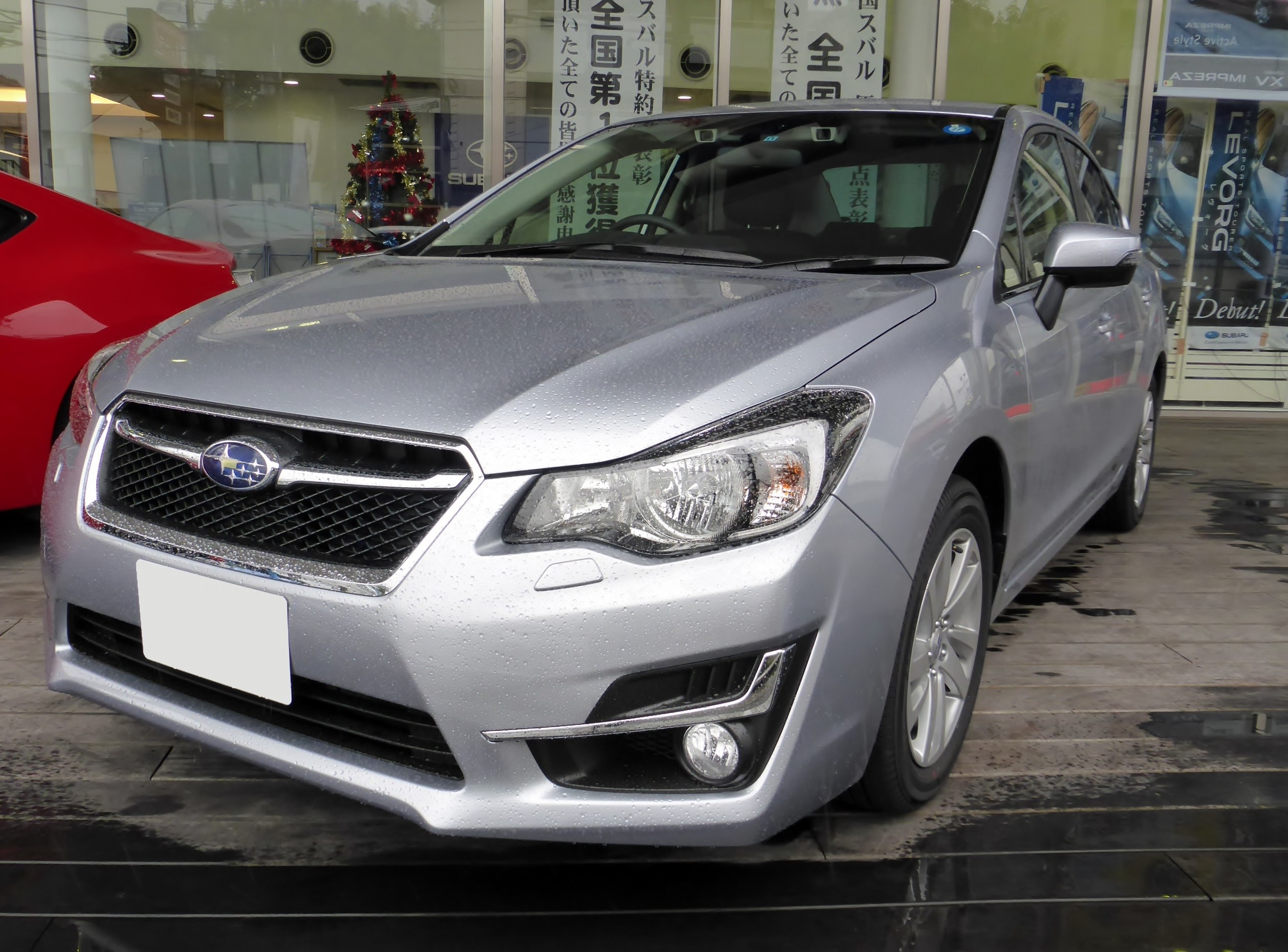 Silberner Wagen vor einem roten Wagen geparkt, mit einer Glaswand, die einen dekorierten Innenraum mit einem Weihnachtsbaum und Lichtern sowie Plakate mit Autos auf der rechten Seite zeigt.