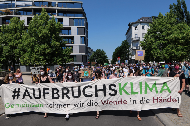 Eine Gruppe von Menschen mit Masken hält ein Banner mit der Aufschrift "Aufbruchsklima" vor einem Gebäude mit Bäumen und einem klaren Himmel, während sie gegen die Klimapolitik der deutschen Regierung protestieren.