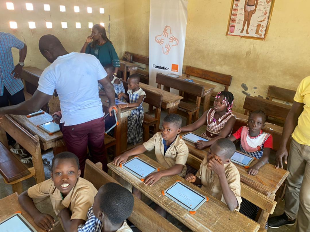 Eine Gruppe von Kindern sitzt an Tischen in einem Klassenzimmer und nutzt Tablets, umgeben von ein paar stehenden Personen, sowie einem Banner und einem Plakat im Hintergrund.