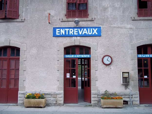 Ein Gebäude mit Türen, Fenstern und einer Uhr an der Wand, mit einer Tafel mit Text, Pflanzen und einer Straße unten.