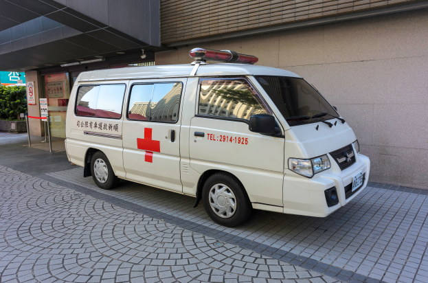 Ein Rettungswagen steht vor einem Geb√§ude mit einer Tafel, umgeben von B√§umen und Pflanzen.