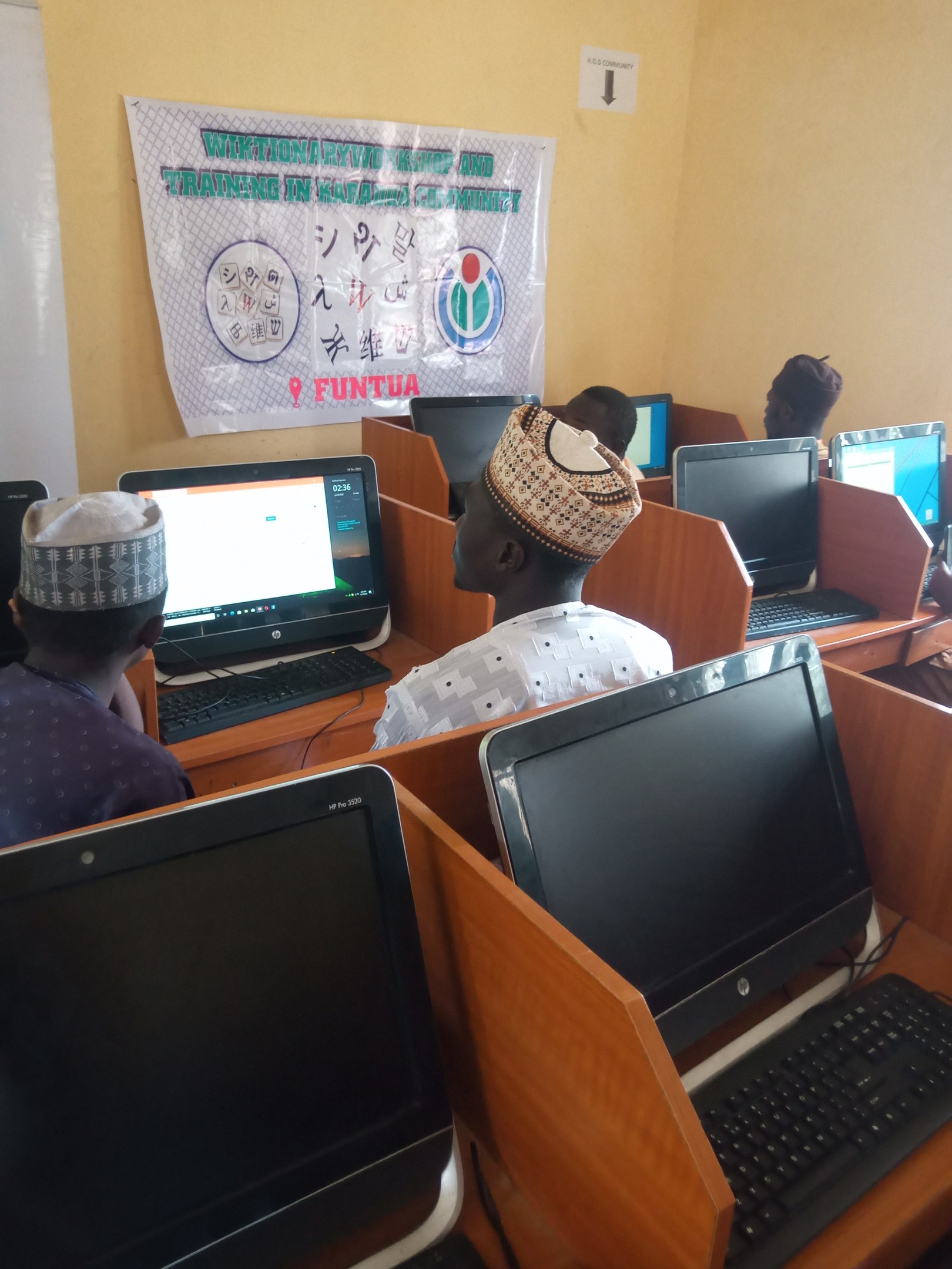 Eine Gruppe von Menschen, die an Tischen mit Computern, Tastaturen und Mäusen sitzen und an einem Workshop zur Computertechnologie teilnehmen, mit einem Plakat im Hintergrund.