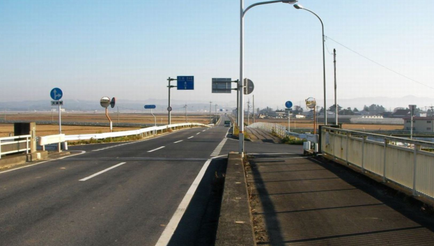Eine Autobahn mit einer Brücke, die Geländer, Laternenmästele und Verkehrszeichen hat, mit Gebäuden, Bäumen, Hügeln und einem klaren blauen Himmel im Hintergrund.
