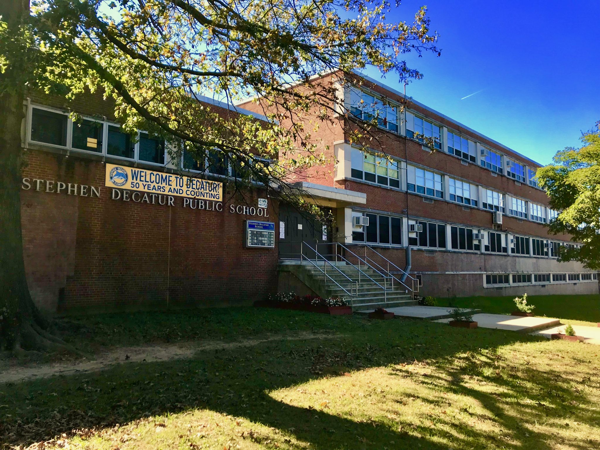 Stephen Decatur Public School-Gebäude mit Fenstern, einer Treppe mit Geländern, einem Schild, Topfpflanzen, Gras, einer Gruppe von Bäumen und einem bewölkten Himmel.