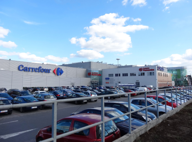 Ein großes Autohaus mit zahlreichen parkenden Autos, umgeben von Gebäuden, Straßenlaternen, Straßenschildern, Motorrädern auf der Straße, Gittern und einem bewölkten Himmel im Hintergrund.