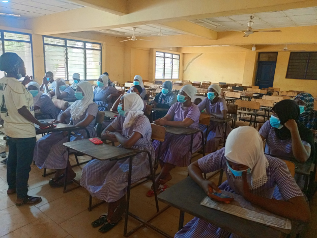 Eine Klasse in Nigeria mit Schülern, die Gesichtsmasken tragen und auf Bänken sitzen oder stehen, mit Büchern und Gegenständen auf Tischen, Fenstern, einer Tür und Deckenlampen und Ventilatoren im Hintergrund.